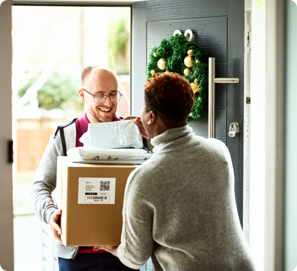 Delivery man standing next to open door with holiday wreath handing woman several packages.